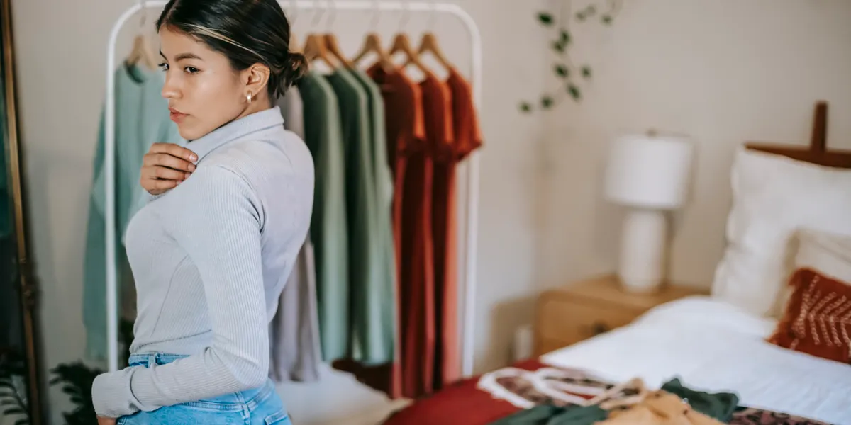 A woman in a pale ribbed turtleneck standing beside an open clothing rail in a bedroom, adjusting her collar